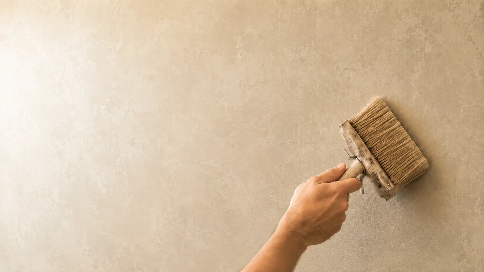 A wide brush laying soft lime wash across a warm, clouded beige wall, showing the hand-applied brushed movement of the finish.