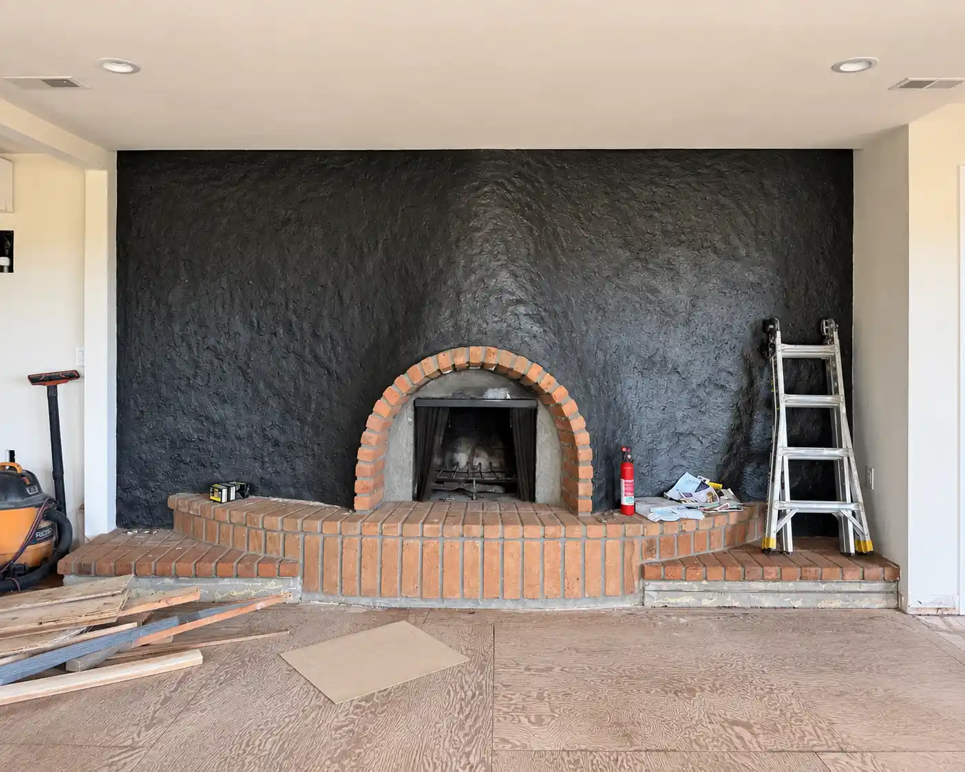 Fireplace before microcement, a black textured wall with a red brick arch and two-level hearth, during renovation in a San Diego, Orange County area home.