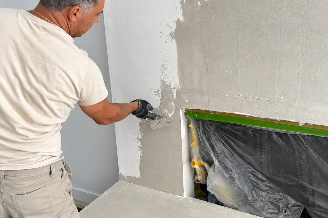 Specialist hand-troweling the plaster surface at the corner beside the fireplace opening.