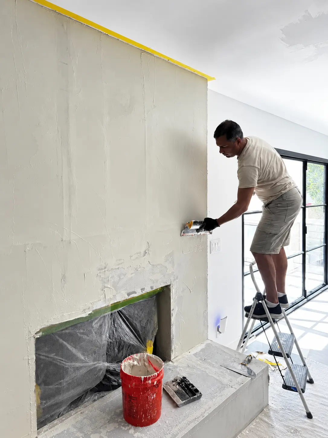 Specialist on a step ladder pulling plaster to a smooth plane on the upper fireplace wall, firebox masked below.