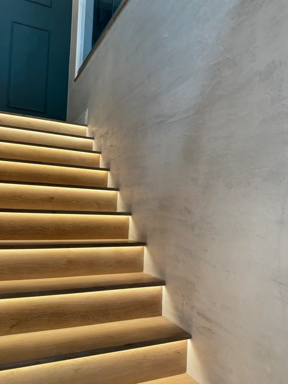 Finished Venetian plaster stairwell in a warm coffee travertine tone beside the lit wood steps, Orange County.